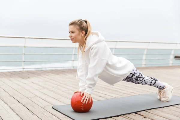 Deniz kenarında çalışırken fitness boll ile fitness mat egzersizleri yapan odaklanmış sportif kadın Görüntü — Stok fotoğraf