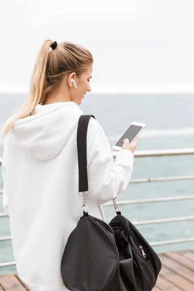 Bild av vacker ung kvinna med hjälp av mobiltelefon och EarPods medan du går nära havet med sportig väska — Stockfoto