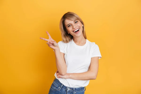 Glückliche junge hübsche blonde Frau posiert isoliert über gelbem Wandhintergrund in weißem lässigem T-Shirt, das Friedensgeste zeigt. — Stockfoto