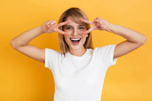 Optimistische emotionele jonge mooie blonde vrouw poseren geïsoleerd over gele muur achtergrond gekleed in witte casual t-shirt tonen vrede gebaar. — Stockfoto