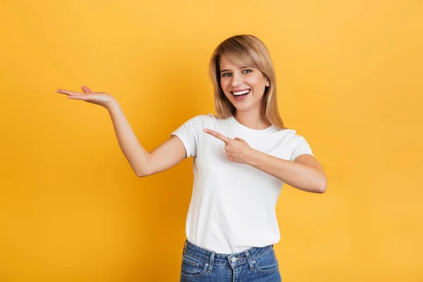 Hermosa feliz joven rubia complacida posando aislada sobre fondo amarillo de la pared vestida con una camiseta casual blanca que apunta al copyspace . — Foto de Stock