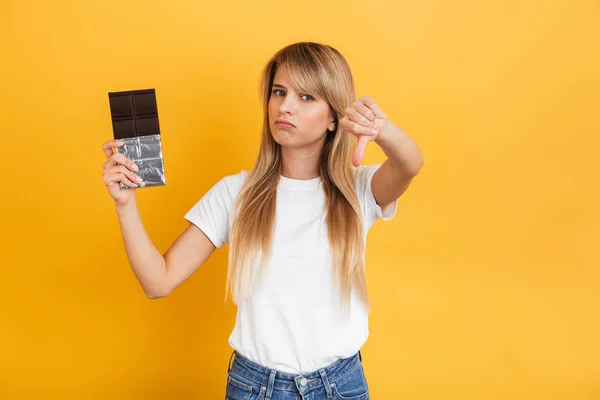 Mulher loira jovem posando isolado sobre fundo de parede amarelo vestido em branco casual t-shirt segurando doces de chocolate mostrando polegares para baixo . — Fotografia de Stock