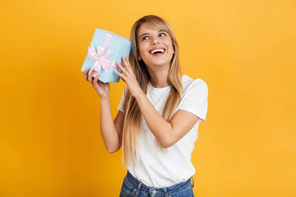 Feliz emocionalmente alegre joven rubia posando aislada sobre fondo amarillo de la pared vestida con una camiseta casual blanca sosteniendo la caja de regalo presente . —  Fotos de Stock