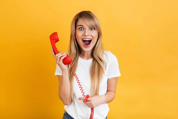 Feliz joven sorprendida rubia posando aislada sobre fondo amarillo de la pared hablando por teléfono rojo retro . — Foto de Stock