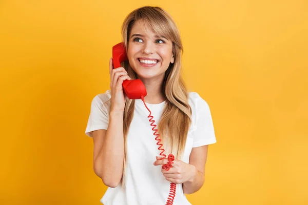 Sonriente emotiva joven rubia feliz posando aislada sobre fondo amarillo de la pared vestida con una camiseta blanca casual hablando por teléfono . — Foto de Stock