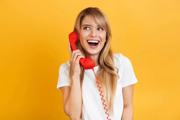 Sonriente emotiva joven rubia feliz posando aislada sobre fondo amarillo de la pared vestida con una camiseta blanca casual hablando por teléfono . — Foto de Stock