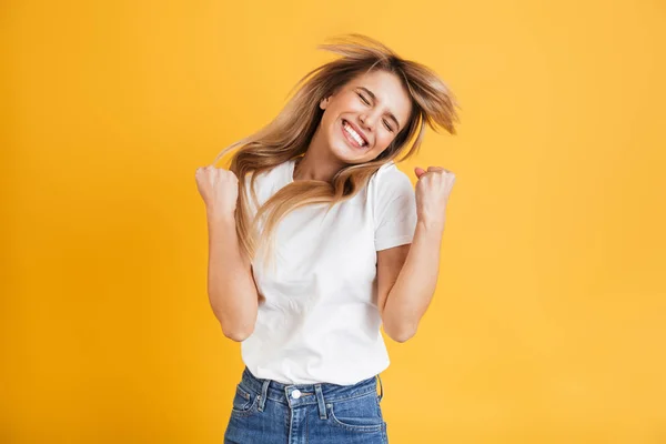 Mulher loira jovem emocional posando isolado sobre fundo de parede amarelo vestido em branco casual t-shirt mostrando gesto vencedor . — Fotografia de Stock