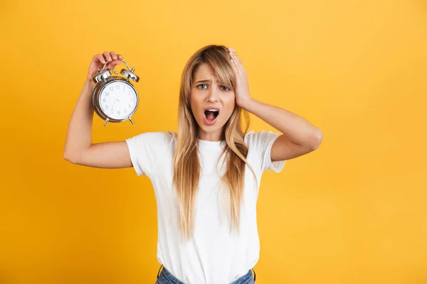 Gritando triste joven rubia posando aislada sobre fondo amarillo de la pared vestida con camiseta casual blanca sosteniendo reloj despertador . — Foto de Stock