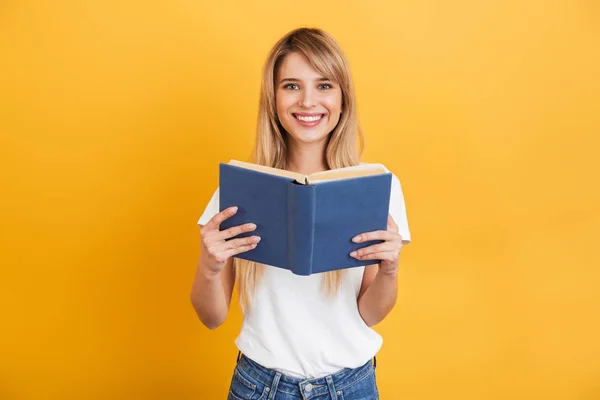 Cheerful emotional young blonde woman posing isolated over yellow wall background dressed in white casual t-shirt holding book reading. — Stock Photo, Image