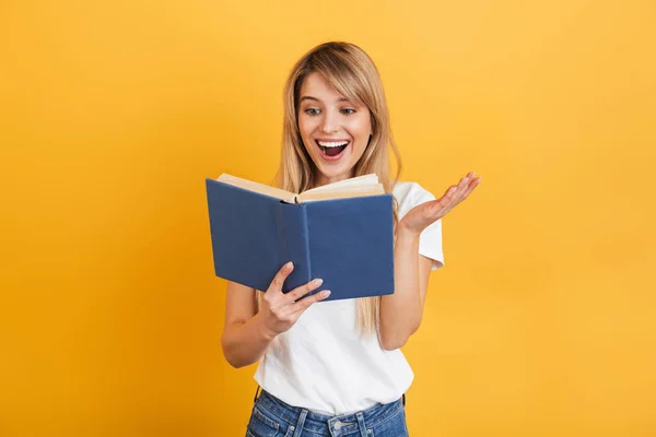 Neşeli duygusal genç sarışın kadın beyaz rahat t-shirt kitap okuma tutan giymiş sarı duvar arka plan üzerinde izole poz. — Stok fotoğraf