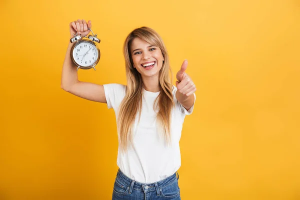 Glad leende ung blond kvinna poserar isolerade över gul vägg bakgrund klädd i vitt casual t-shirt hålla väckarklocka visar tummen upp. — Stockfoto