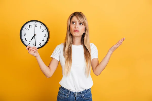 Thinking young blonde woman posing isolated over yellow wall background dressed in white casual t-shirt holding clock. — Stock Photo, Image