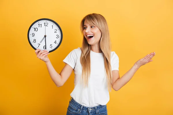 Vrolijke jonge blonde vrouw poseren geïsoleerd over gele muur achtergrond gekleed in witte casual t-shirt Holding klok. — Stockfoto