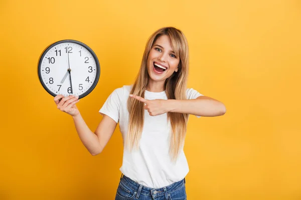 Cheery young blonde woman posing isolated over yellow wall background dressed in white casual t-shirt holding clock pointing. — Stock Photo, Image