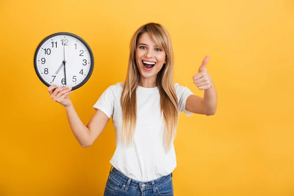 Happy young blonde woman posing isolated over yellow wall background dressed in white casual t-shirt holding clock shows thumbs up . — Stok Foto