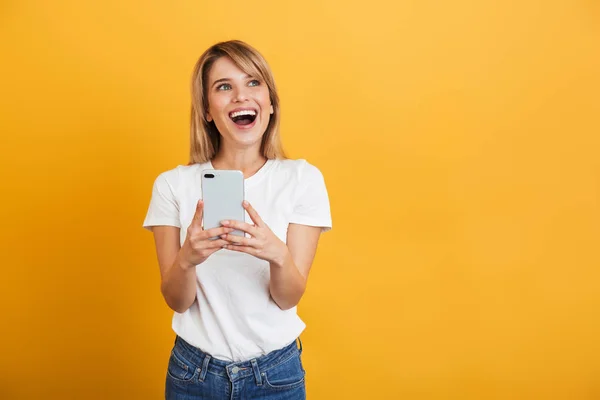 Happy emotional young blonde woman posing isolated over yellow wall background dressed in white casual t-shirt using mobile phone. — Stock Photo, Image