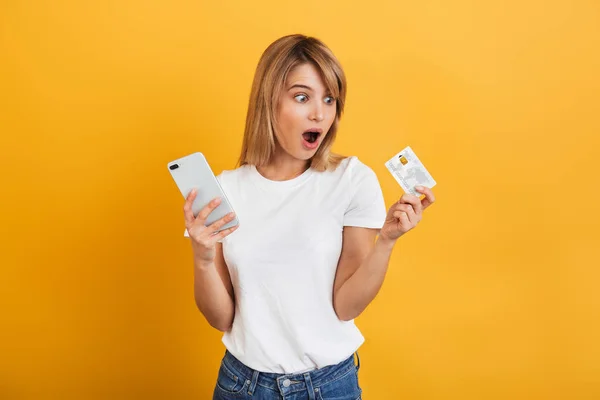 Chockad förvånad ung blond kvinna poserar isolerade över gul vägg bakgrund klädd i vitt casual t-shirt med hjälp av mobiltelefon Holding kreditkort. — Stockfoto