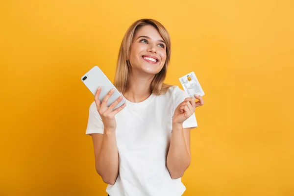 Happy young blonde woman posing isolated over yellow wall background dressed in white casual t-shirt using mobile phone holding credit card. — Stock Photo, Image