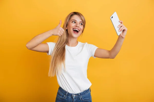 Gelukkig jonge blonde vrouw poseren geïsoleerd over gele muur achtergrond gekleed in witte casual t-shirt met behulp van mobiele telefoon neem een selfie. — Stockfoto