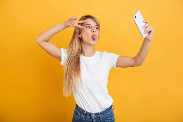 Szczęśliwy młody Blondynka kobieta pozowanie na białym tle na żółtym ścianie tło ubrany w biały casual t-shirt za pomocą telefonu komórkowego zrobić selfie. — Zdjęcie stockowe