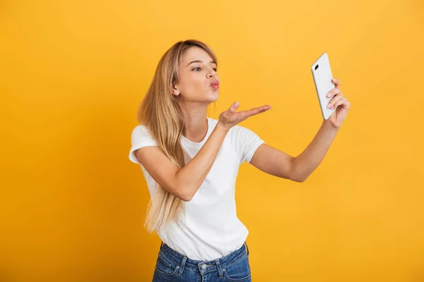 Lycklig ung blond kvinna poserar isolerade över gul vägg bakgrund klädd i vit casual t-shirt med mobiltelefon ta en selfie. — Stockfoto