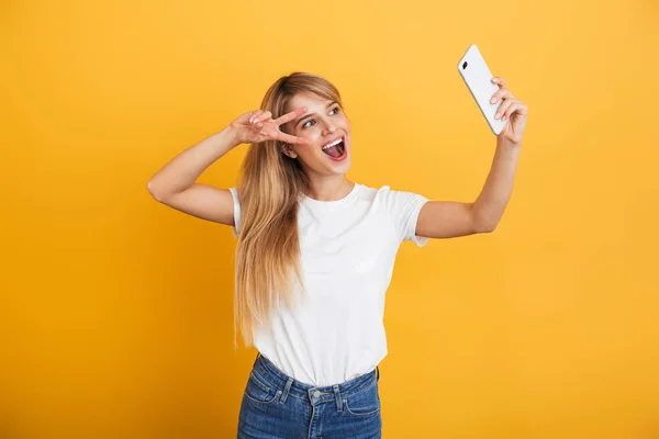 Happy young blonde woman posing isolated over yellow wall background dressed in white casual t-shirt using mobile phone take a selfie. — Stock Photo, Image