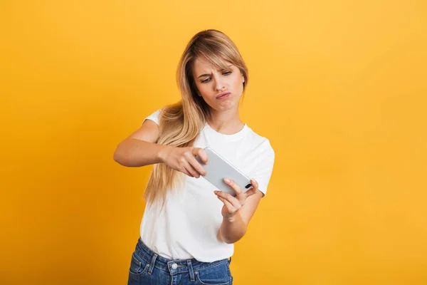 Nervös ung blond kvinna poserar isolerade över gul vägg bakgrund klädd i vit casual t-shirt med mobiltelefon spela spel. — Stockfoto