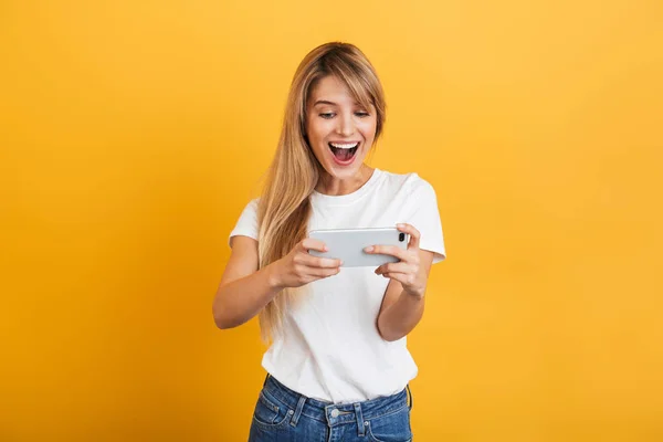Gelukkige emotionele jonge blonde vrouw poseren geïsoleerd over gele muur achtergrond gekleed in witte casual t-shirt met behulp van mobiele telefoon spelen spelletjes. — Stockfoto