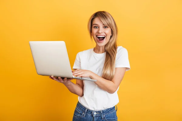Positivo otimista emocional jovem loira mulher posando isolado sobre amarelo parede fundo vestido em branco casual t-shirt usando laptop computador . — Fotografia de Stock
