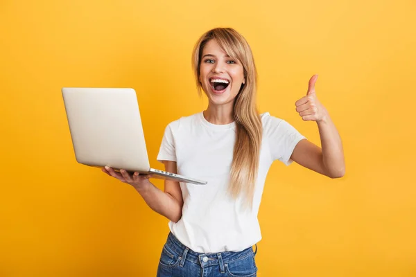 Positiva optimista joven rubia emocional posando aislada sobre fondo amarillo de la pared vestida con una camiseta casual blanca usando computadora portátil . — Foto de Stock