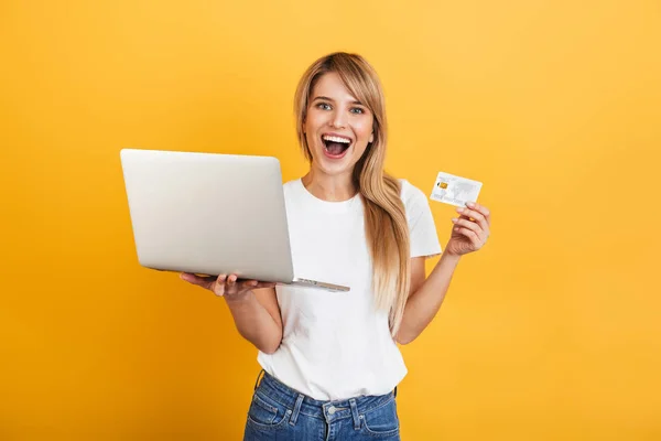ポジティブ楽観的な感情的な若いブロンドの女性は、ラップトップコンピュータを使用して白いカジュアルなTシャツを着て黄色の壁の背景の上に孤立したポーズ. — ストック写真