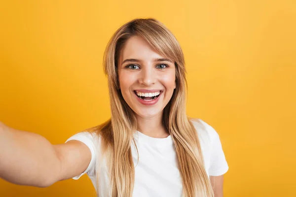 Щаслива позитивна приємна молода блондинка позує ізольована на жовтому фоні стіни, одягнена в білу повсякденну футболку візьміть селфі на камеру . — стокове фото