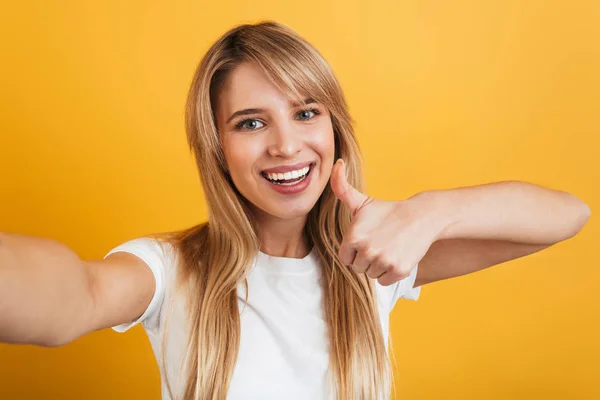 Feliz positivo complacido joven rubia posando aislado sobre fondo amarillo de la pared vestida con blanco casual camiseta tomar selfie por cámara . — Foto de Stock