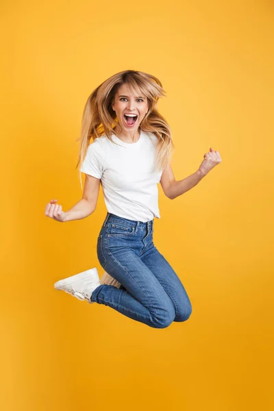 Emotionale Schreie positive junge blonde Frau springt isoliert über gelbe Wand Hintergrund gekleidet in weißen lässigen T-Shirt machen Siegergeste. — Stockfoto