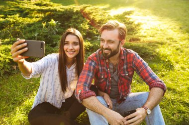 Yeşil parkta dinlenirken gülümseyen çift erkek ve kadının cep telefonundan selfie fotoğrafı çekmesi