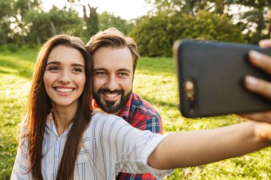 Yeşil parkta dinlenirken cep telefonundan selfie fotoğrafı çeken mutlu çift erkek ve kadının görüntüsü