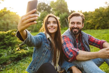 Yeşil parkta dinlenirken sevimli çift erkek ve kadının cep telefonundan selfie fotoğrafı çekmesi görüntüsü