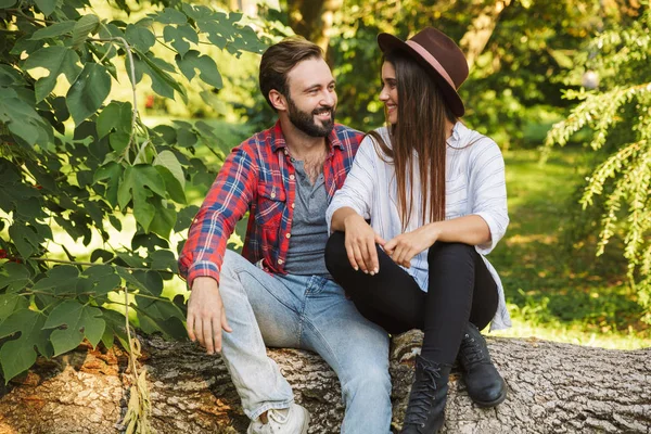Изображение привлекательной пары мужчины и женщины, улыбающихся сидя на дереве в зеленом парке — стоковое фото
