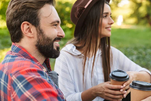 Beeld van aantrekkelijke paar man en vrouw drinken afhaalkoffie terwijl rusten in groen park — Stockfoto