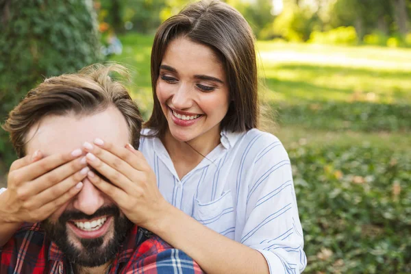 Image of beautiful woman covering man\'s eyes while resting in green park
