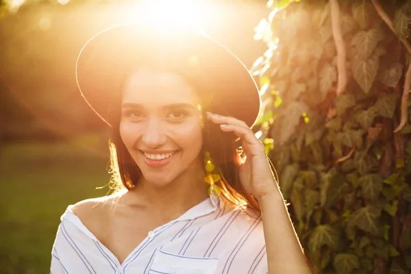 Foto de una linda mujer joven con un sombrero elegante sonriendo y caminando en el parque verde — Foto de Stock