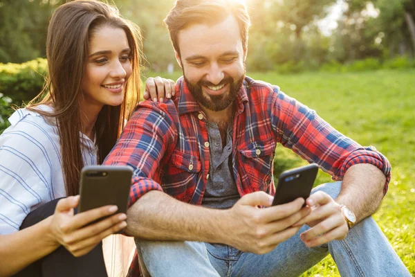 Imagen de pareja atractiva hombre y mujer usando teléfonos celulares mientras descansan en el parque verde — Foto de Stock