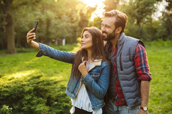 Yeşil parkta yürürken sevimli çift erkek ve kadının cep telefonundan selfie fotoğrafı çekmesi görüntüsü
