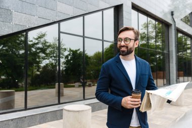 Gözlük takan mutlu işadamının kâğıt bardaktan kahve içerken ve dışarıda gazete okumaportresi