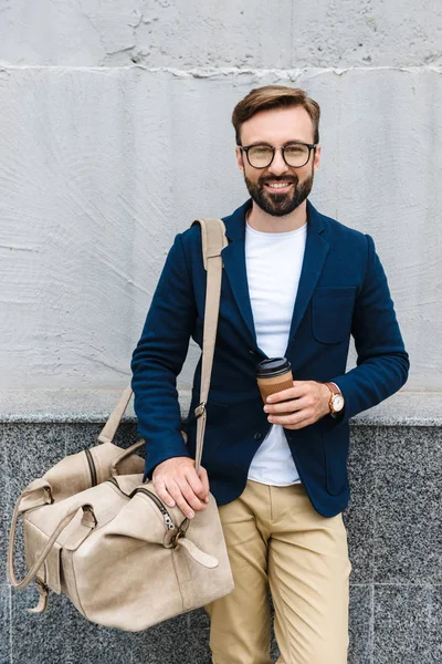 Retrato de un hombre de negocios barbudo con anteojos sosteniendo una taza de papel y una bolsa de transporte — Foto de Stock