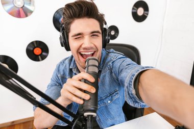Yakışıklı genç adam radyo sunucusu mikrofon ve ses ekipmanları ile çalışma alanında kamera ile selfie çekmek.