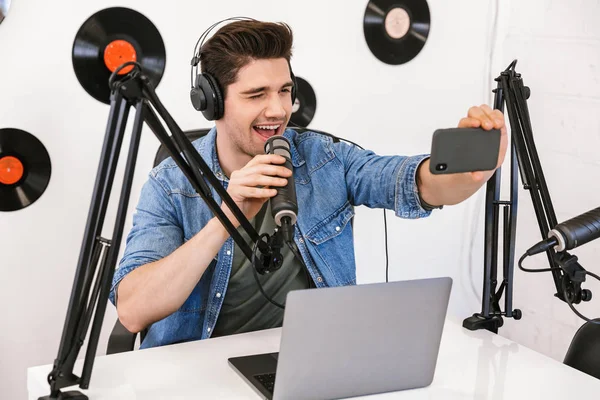 Schöner junger Mann Radiomoderator am Arbeitsplatz mit Mikrofon und Tonanlage macht Selfie mit dem Handy. — Stockfoto