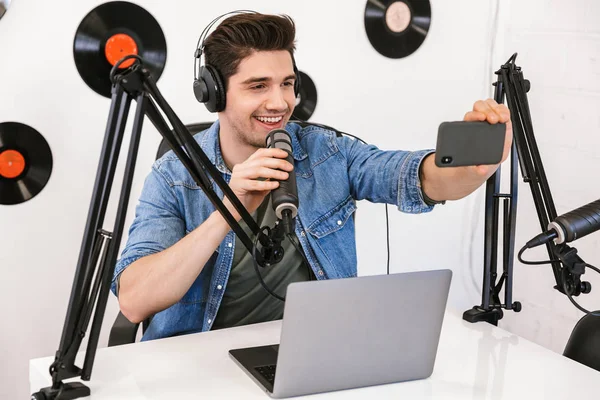 Bel giovane conduttore radiofonico nello spazio di lavoro con microfono e apparecchiature audio scattare selfie dal telefono cellulare . — Foto Stock