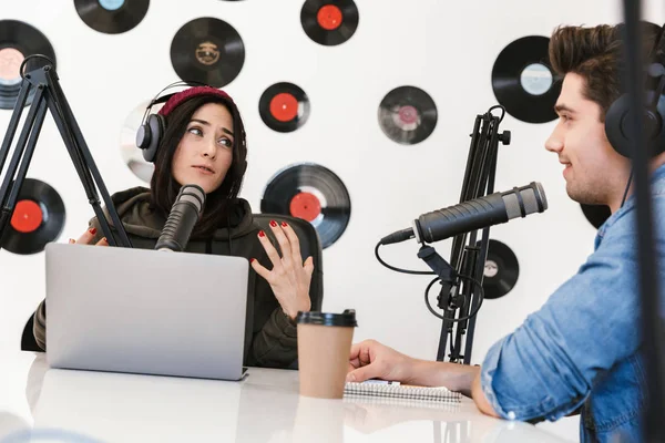 Jovem apresentador de rádio com colega mulher no espaço de trabalho com microfone e equipamentos de som conversando uns com os outros . — Fotografia de Stock