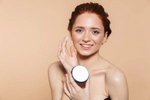 Increíble joven pelirroja posando aislada sobre fondo de pared beige cuidar su piel con crema . —  Fotos de Stock